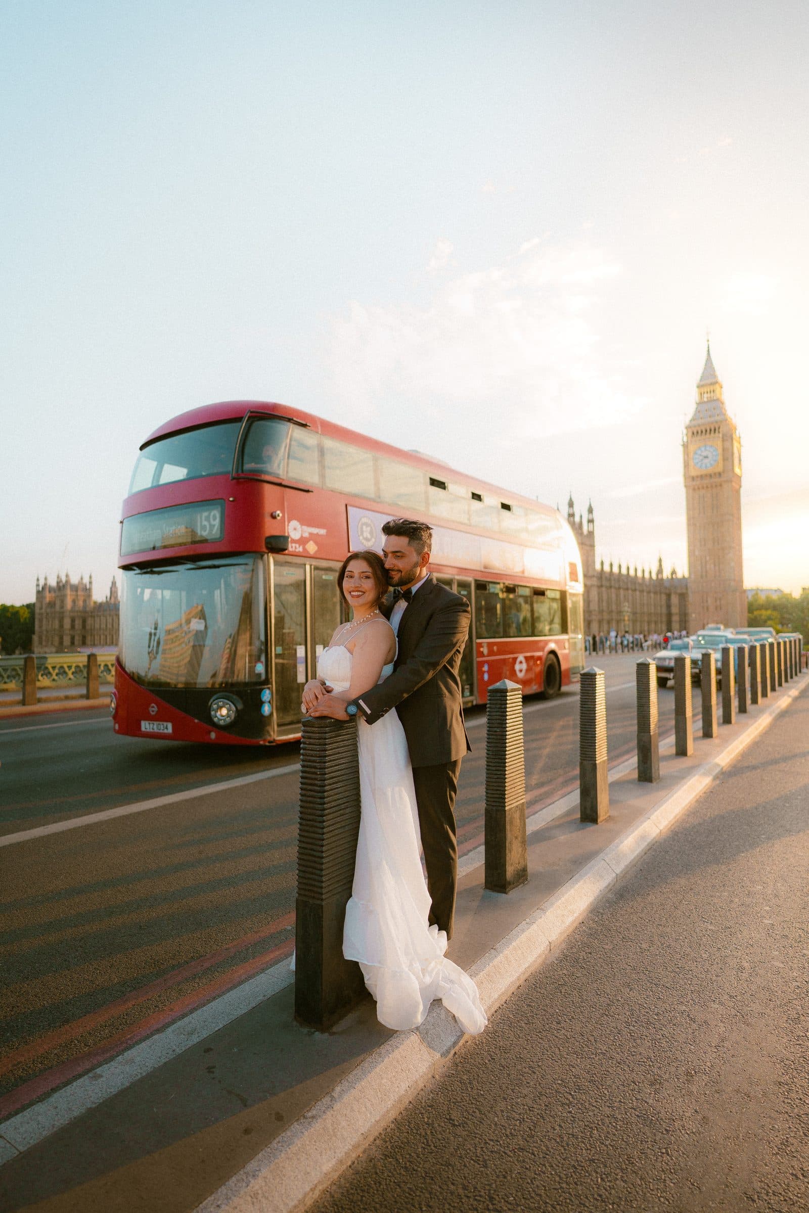 A couple in wedding clothes on Westminster Bridge as a red double-decker bus passes
