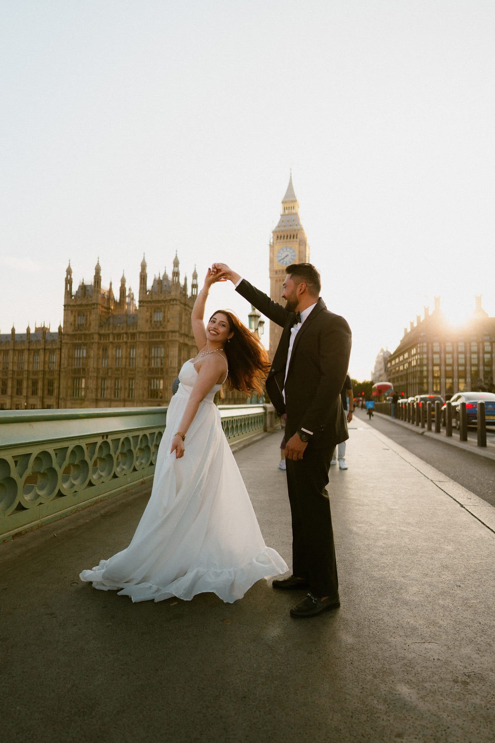 A couple mid-twirl on Westminster Bridge at golden hour, her white dress flaring in front of Big Ben